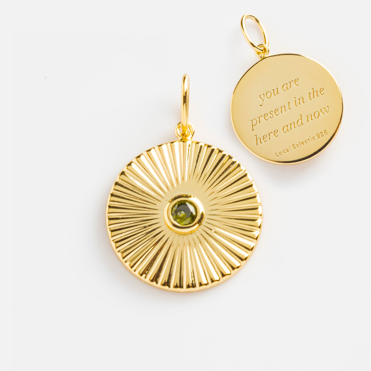 Two round 14k gold plated pendants are shown: one features a sunburst pattern with a green gemstone center, and the other is the Mantra Empowerment Coin Charm engraved with “you are present in the here and now.”.