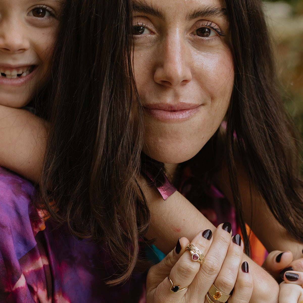 A woman and child in colorful tie-dye embrace, both displaying painted nails and rings, including the Solid Gold Citrine Bold Fluted Cigar Band. The scene is set against a backdrop of blurred greenery as they smile warmly.