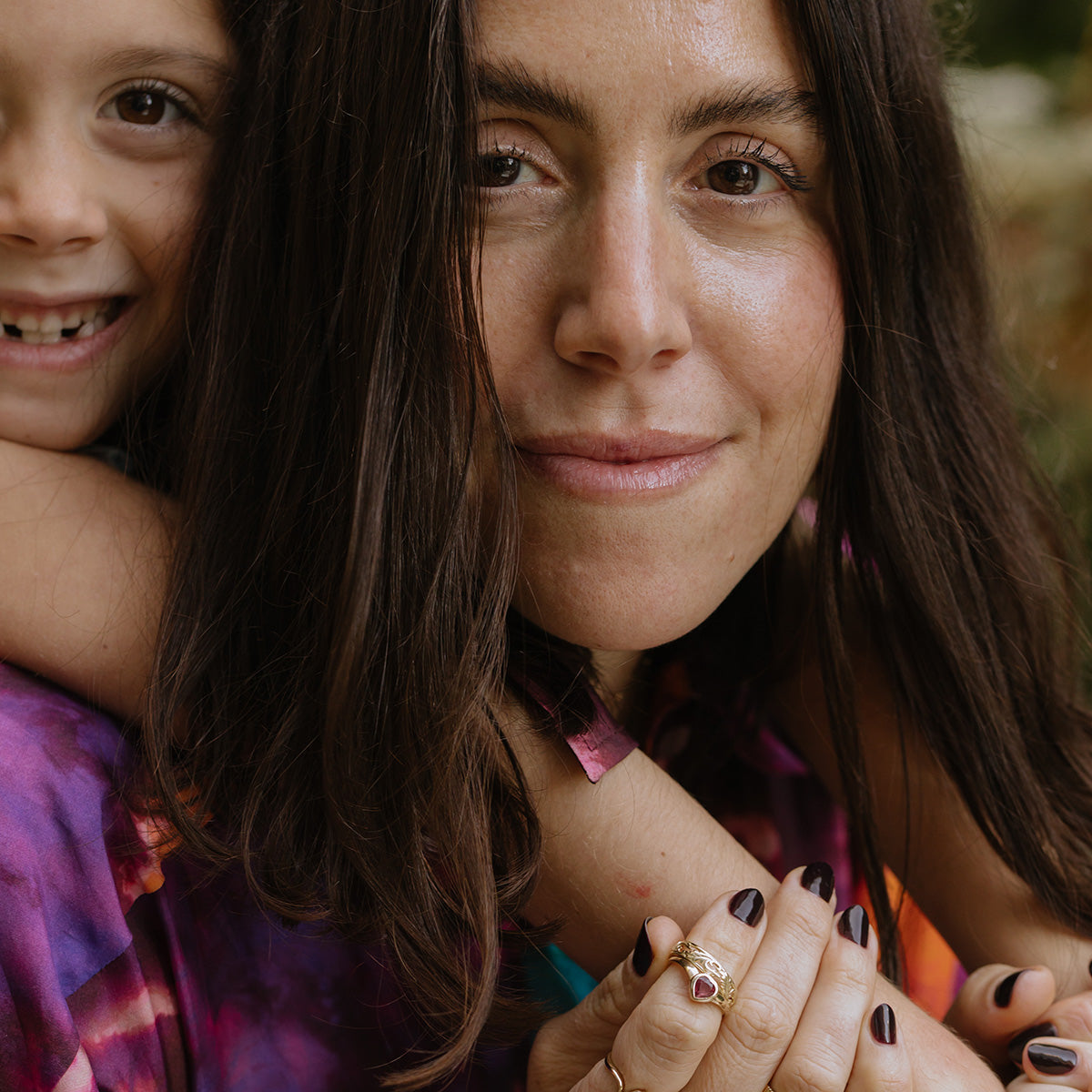 A woman with long dark hair smiles at the camera as a child hugs her from behind, both in colorful clothes. Her hand, with dark nail polish and a Solid Gold Bezel Set Ruby Heart Ring, is visible in the foreground.