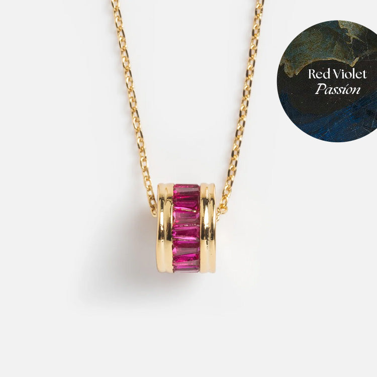 The Color Theory Charm Necklace features a gold plated round pendant with vertical red violet CZ stones. A circular label reading Red Violet Passion sits on a dark, artistic background. The perfect vibrant accessory.