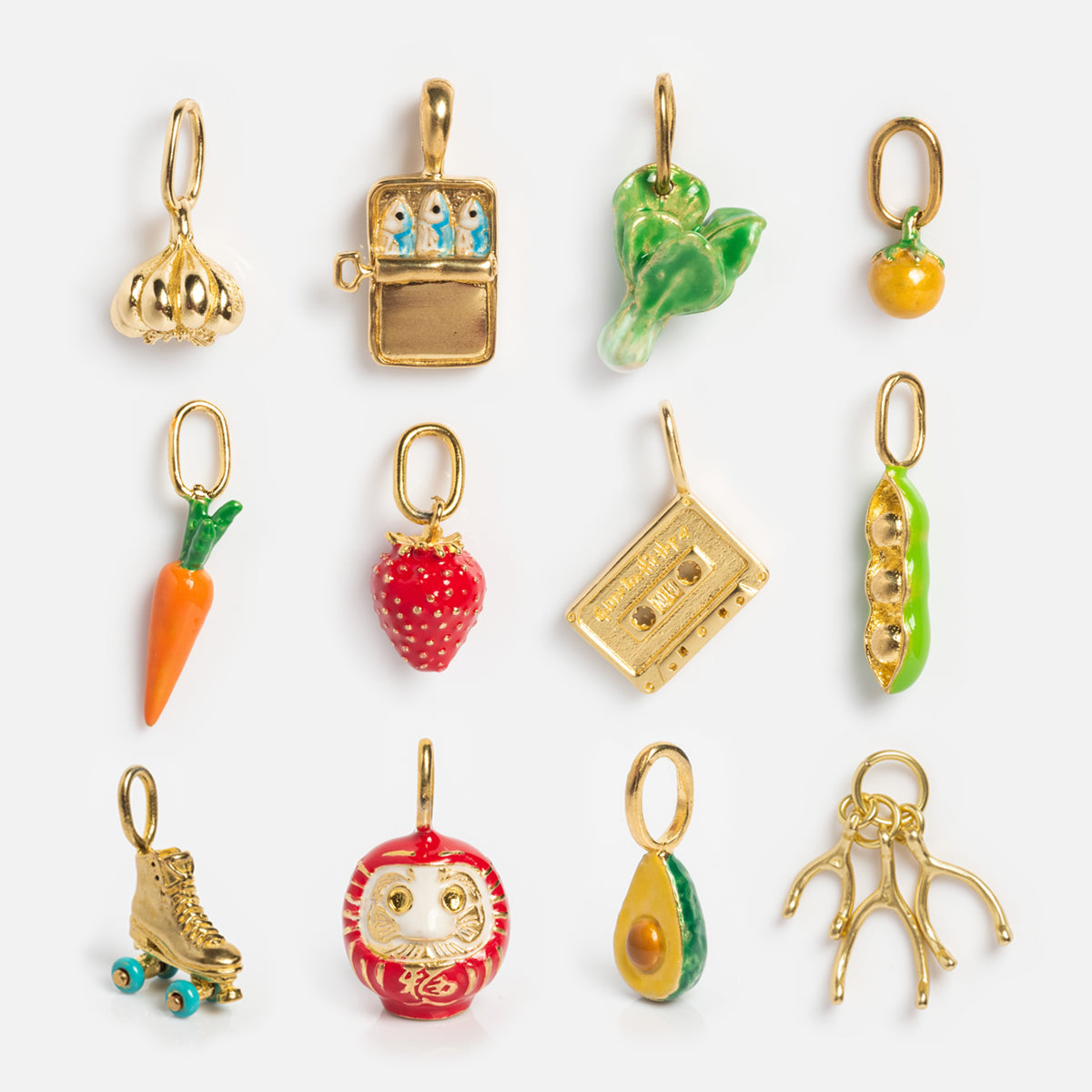 Charms by Satomi Studio features twelve small enamel jewelry pieces arranged in rows, including necklace charms shaped like garlic, sardines in a tin, lettuce, orange, carrot, strawberry, cassette tape, peas, roller skate, daruma doll, avocado, and wishbone.