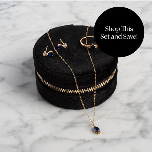 The Solid Gold 2025 September Capsule Blue Sapphire Statement Set, featuring earrings, ring, and necklace in a round black velvet zip case, comes with a free gift. Overlay: Shop This Set and Save! on a marble surface.