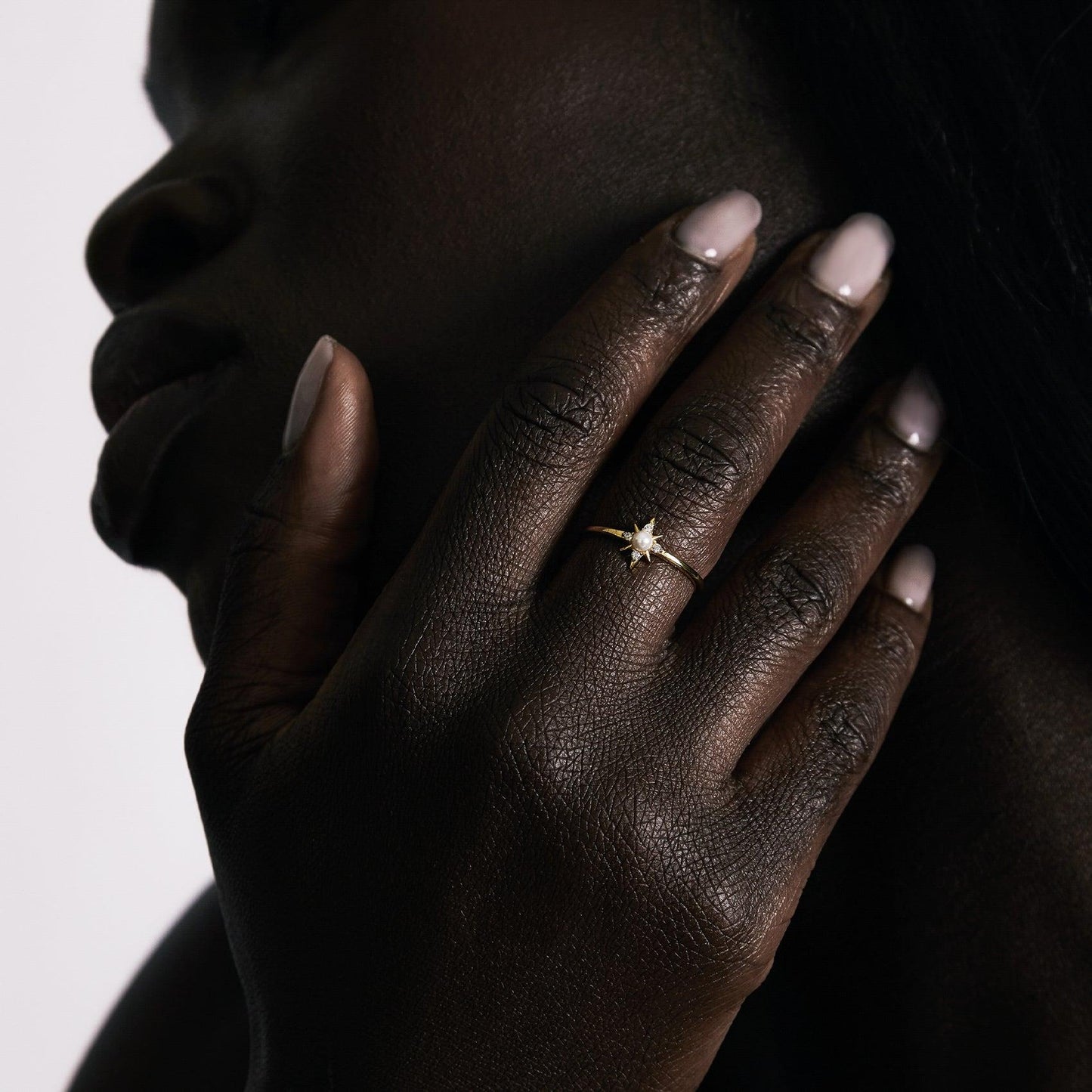 A person with dark skin touches their face, showing off the Celestina Pearl Ring in 14k yellow gold vermeil. Their neatly manicured nude nails and a softly lit background complement the ring’s subtle elegance.