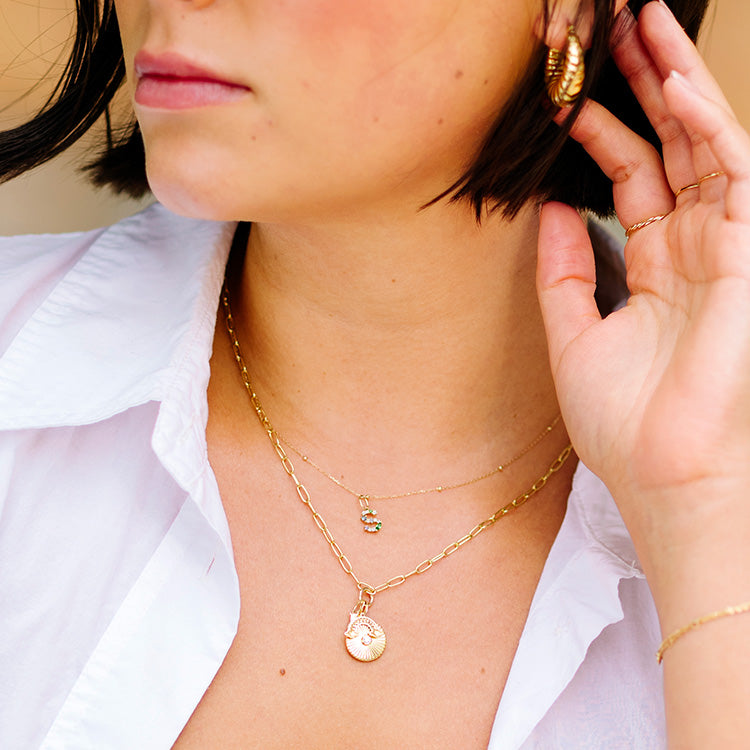 A woman in a white shirt showcases layered gold necklaces—one featuring the Solid Gold Gemstone Letter Charm and another with colorful beads—plus gold hoop earrings, while her hand touches her short dark hair.