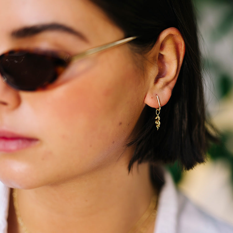 A woman with short dark hair wears tortoiseshell sunglasses and a gold dangling earring, the Solid Gold Nature Charm, shaped like an animal. The softly blurred background features hints of greenery.