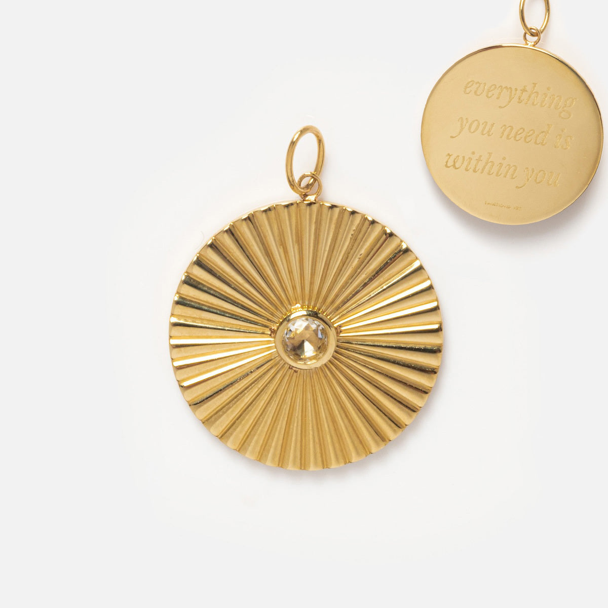 The Solid Gold Coin Charm, a round pendant with radiating grooves and a central gemstone, is displayed beside another Local Eclectic exclusive engraved with everything you need is within you, both set against a light background.