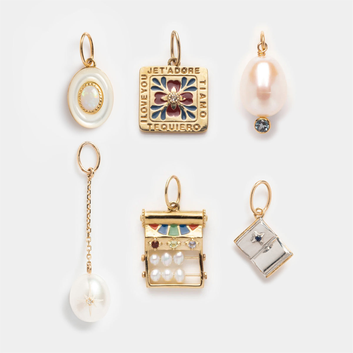 Six Solid Gold Statement Charms are arranged on a white background, featuring unique shapes: a pearl and blue topaz charm, an oval with a gem, colorful love phrase square, globe, jeweled bar, and a small envelope.