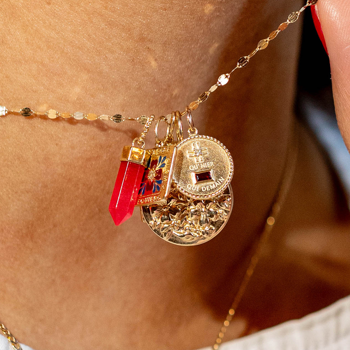 A close-up of the Solid Gold Coin Charm—a gold chain with four pendants including a red crystal, colorful enamel square, textured gold coin, and engraved disc—rests against skin. This exclusive piece is from Local Eclectic.
