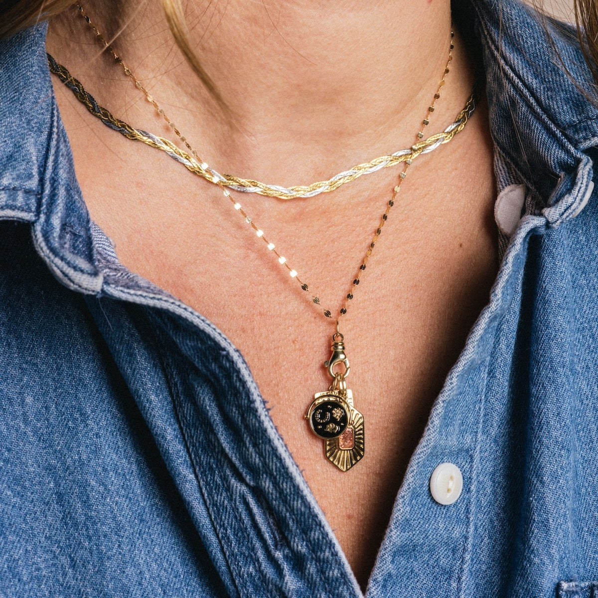 A person wearing a blue denim shirt with two layered gold necklaces: one is a simple chain, and the other features charms, including the Solid Gold Coin Charm and a textured leaf-shaped charm.