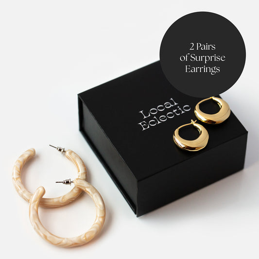 A black jewelry box labeled Local Eclectic holds two pairs of Surprise Box! All Ears earrings—a gold hoop pair and a light marbled designer pair. A circle on the box reads: 2 Pairs of Surprise Box earrings.
