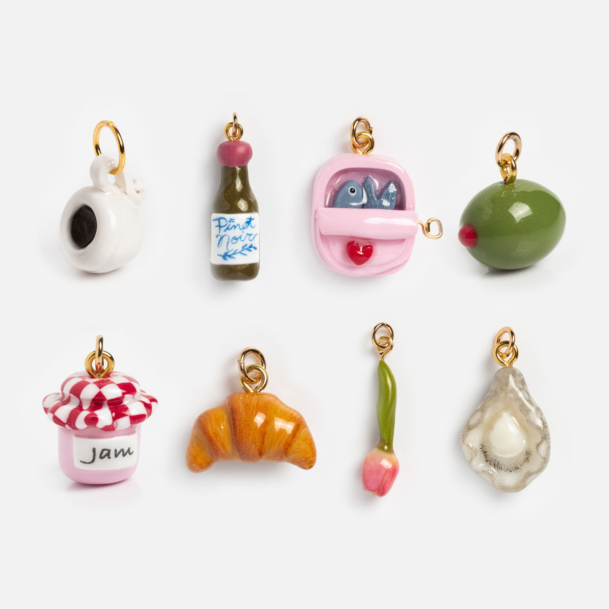 Charms by Warm Clay Co: a set of eight small, colorful gold-plated clay charms shaped like a washing machine, wine bottle, canned fish, olive, jam jar, croissant, tulip, and oyster—each with a gold loop for attaching to necklaces.