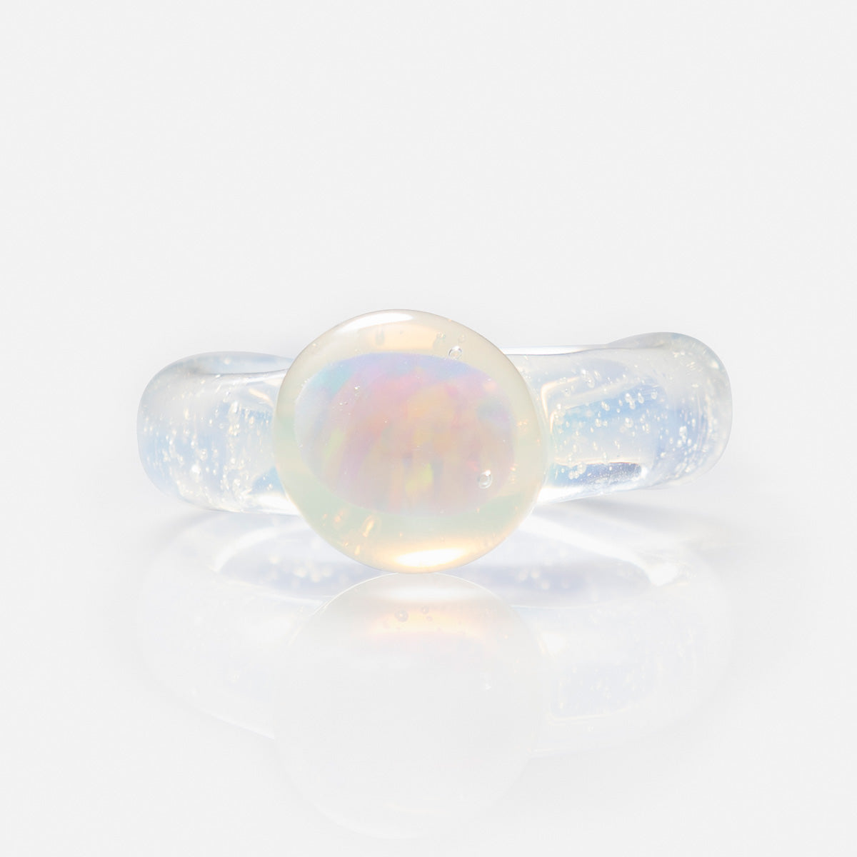 The Small Opal Glass Ring is a translucent, handmade piece with a clear, rounded borosilicate glass band and an oval opal displaying iridescent pink, yellow, and green hues against a white background.