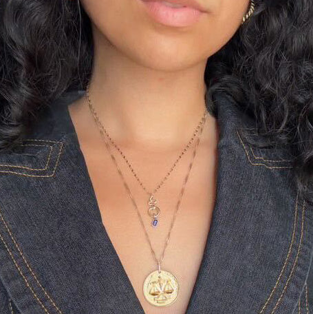 A woman with curly hair wears a denim top and layers two gold necklaces—a vintage charm necklace with a blue stone and the Vintage Inspired Zodiac Coin Necklace.