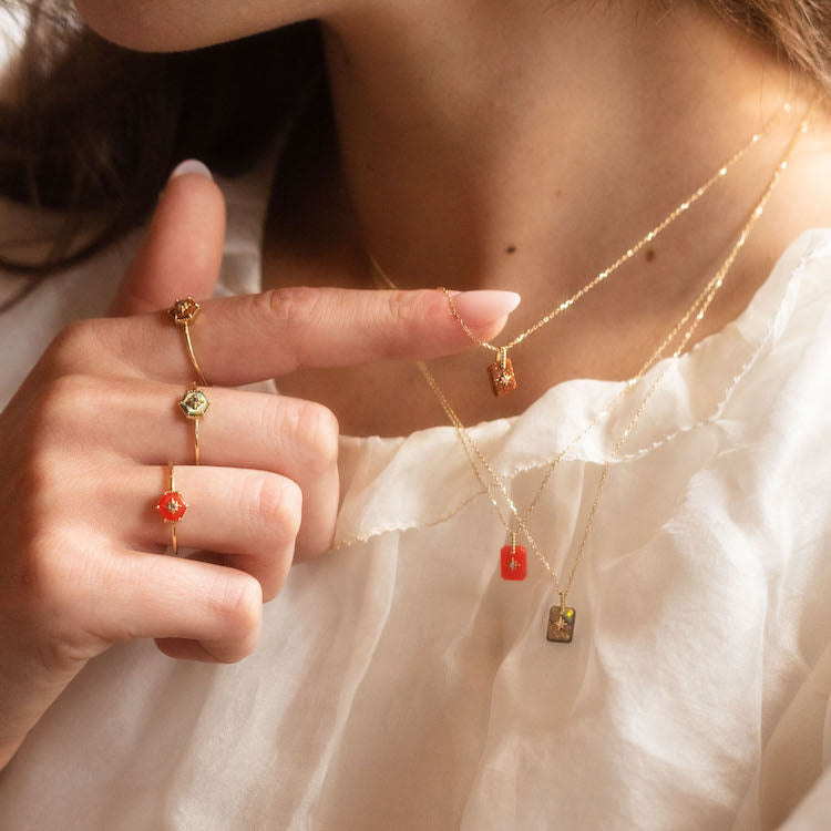 A woman in a white blouse wears gold necklaces and rings, including the Solid Gold Crystal Ring for Manifestation Goldstone (size 7). Her hand, with a yellow gold band, points to a pendant featuring red, orange, green stones and Iolite.