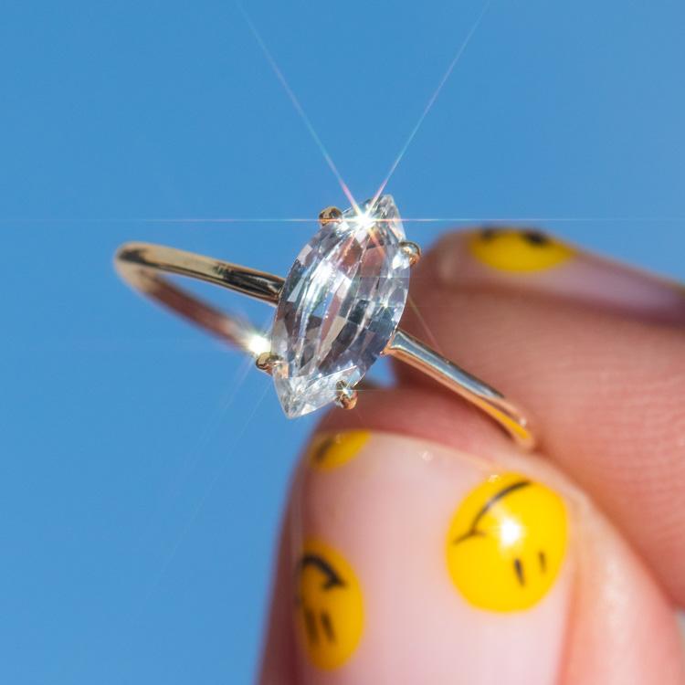 A hand with yellow smiley face nail art holds the Solid Gold Gemstone Marquise Ring in White Topaz, featuring a sparkling clear gemstone, against a bright blue sky.