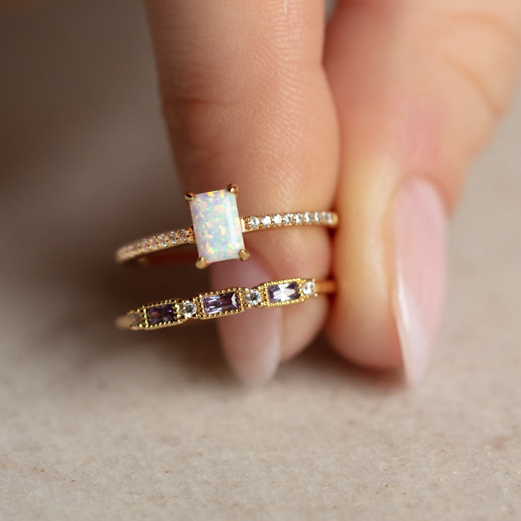 A manicured hand holds two gold rings: the Hopeful Opal Baguette Ring with a luminous opal and small diamonds, and another featuring three purple rectangular stones with detailed yellow gold pavé accents.