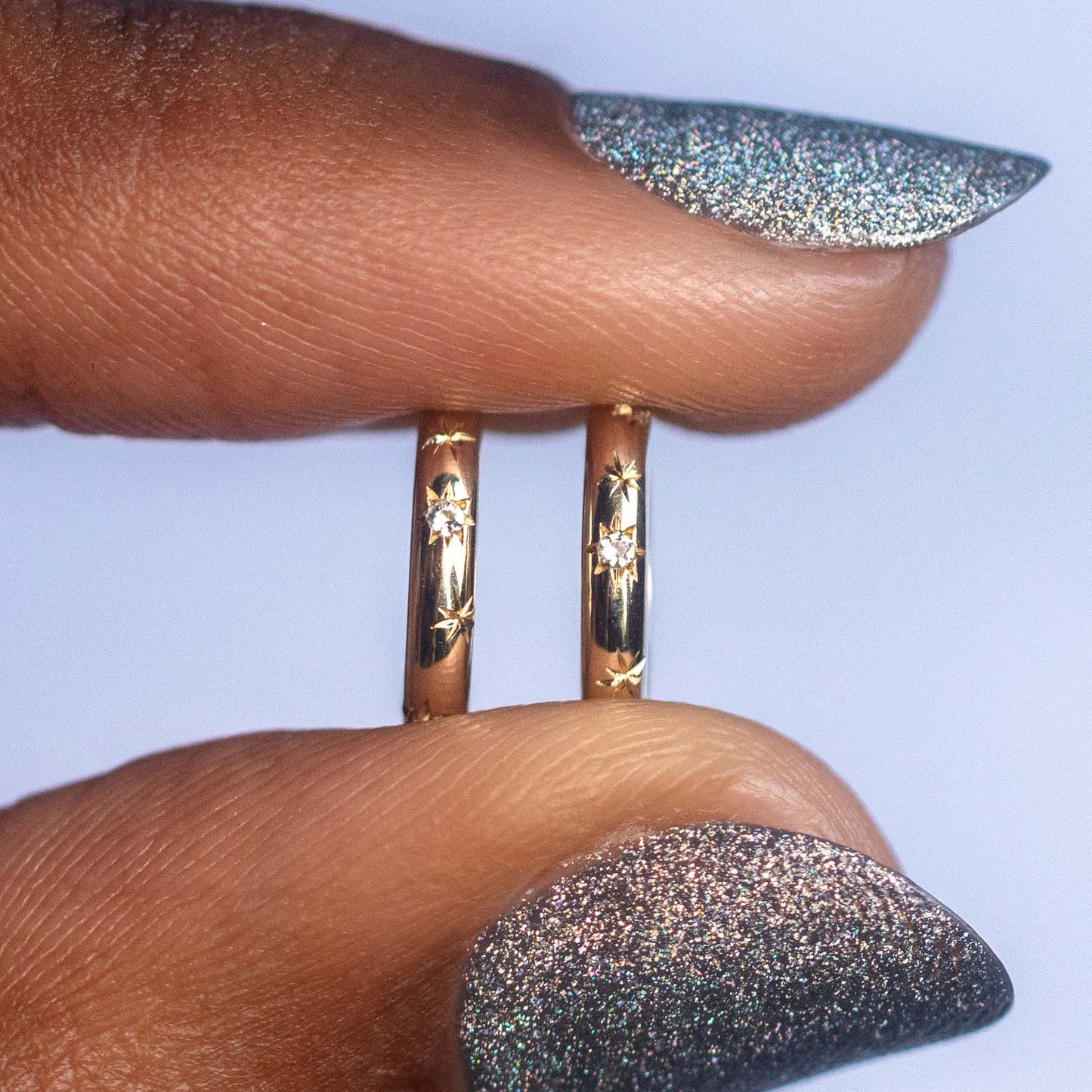 A close-up of two fingers holding Solid Gold Starry White Sapphire Huggies with engraved star designs. Long nails painted with shiny silver glitter polish enhance the celestial allure of these enchanting earrings.