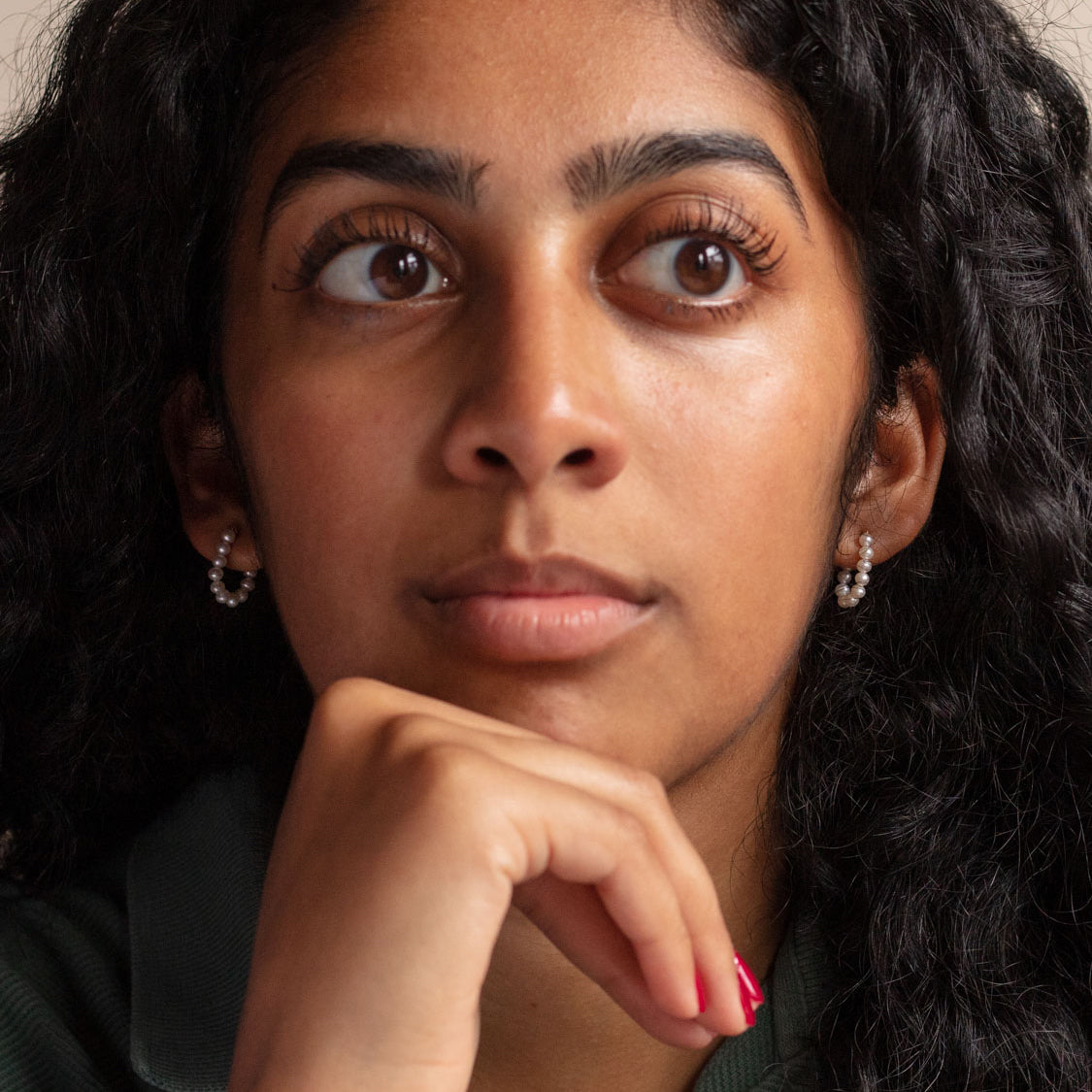 A person with curly dark hair, wearing Solid Gold Pearl Strand Hoops, looks thoughtfully upward with their hand resting near their chin.