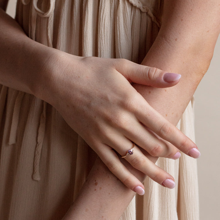 A person with light skin and manicured nails wears the 14k Amethyst and White Topaz Ring Sample Size 7 on their left hand, gently resting it against their arm. They are dressed in a cream-colored, sleeveless dress.