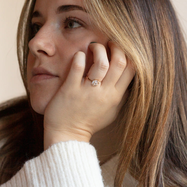 A woman with light brown hair, wearing a cream sweater and the 14k Vintage Inspired Art Deco Engagement Ring, rests her chin on her hand and gazes thoughtfully into the distance.
