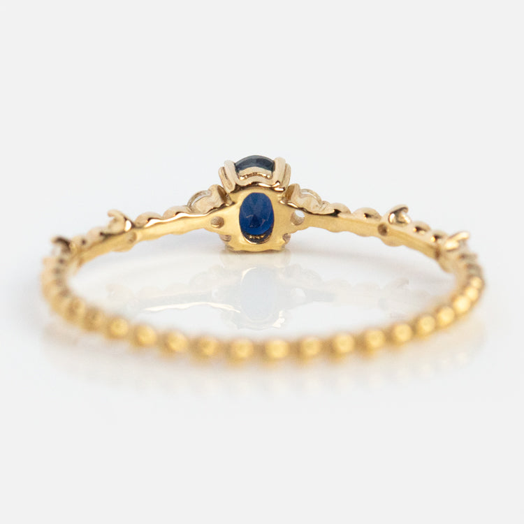 The Solid Gold Something Blue Sapphire Ring features a delicate 9k gold beaded band and an oval blue sapphire in a decorative crown-like prong setting, shown against a white background.