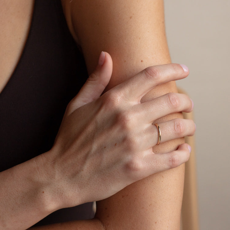 A person’s left hand gently holds their right upper arm, wearing the 14k Delicate Yellow Gold Wedding Band crafted from recycled solid gold. They have on a sleeveless dark top, set against a neutral background.