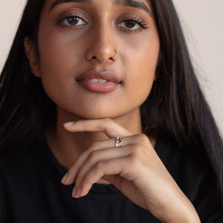 A woman with long dark hair and a nose ring gazes forward calmly, resting her chin on her hand. She wears a black top and the Simple Star Set Diamond Band in yellow gold vermeil on her finger.