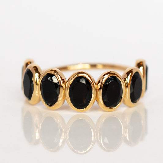 The Violeta Black Onyx Ring showcases seven oval black onyx stones set in smooth 14k yellow gold vermeil bezels, elegantly displayed on a reflective white surface.