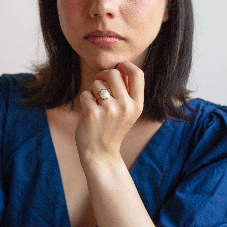 A woman in a deep blue dress rests her chin on her hand, showcasing the Simple Semi Precious Pearl Ring—a gold vermeil ring with a large white pearl—on her finger. Only her lower face and upper torso appear.