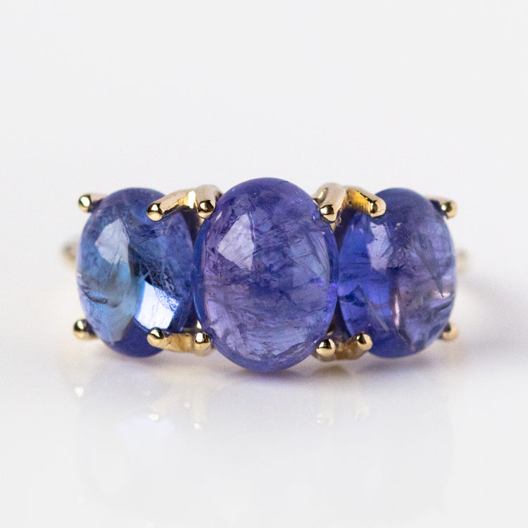 The Sweetest Gumdrop Ring is a yellow gold ring with three large oval-cut natural blue-violet gemstones, each secured by four prongs, shown on a white reflective surface.
