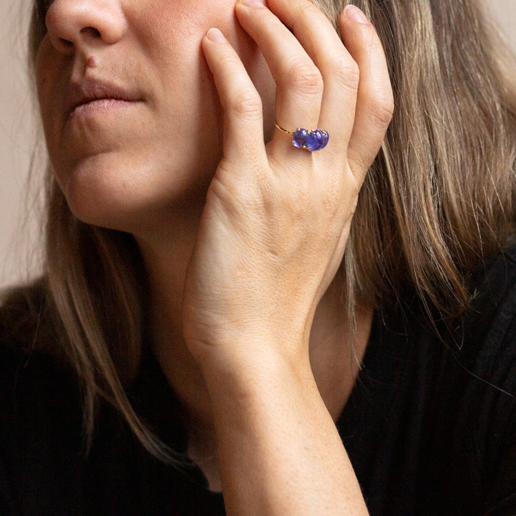 A person with long, light brown hair and a thoughtful expression rests their chin on their hand, wearing the Sweetest Gumdrop Ring in yellow gold set with three purple stones. They are dressed in a black top.