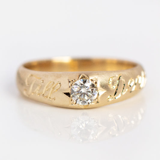 The Till Death Wedding Ring is crafted in 14k solid yellow gold with a round old mine cut diamond set in a star-shaped setting and Till Death engraved in cursive on the band. Photographed on a white background.