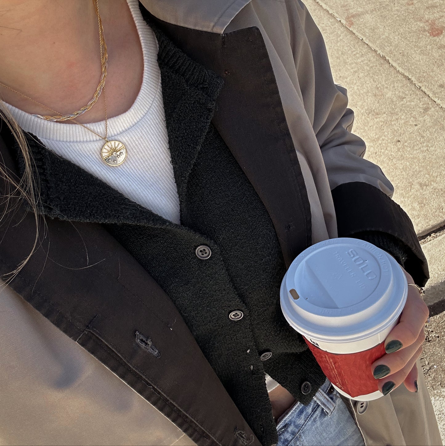 A person wearing layered jackets, a white ribbed shirt, and a Solid Gold Mixed Metal Braided Necklace holds a red disposable coffee cup with a white lid while standing on a sunlit sidewalk.