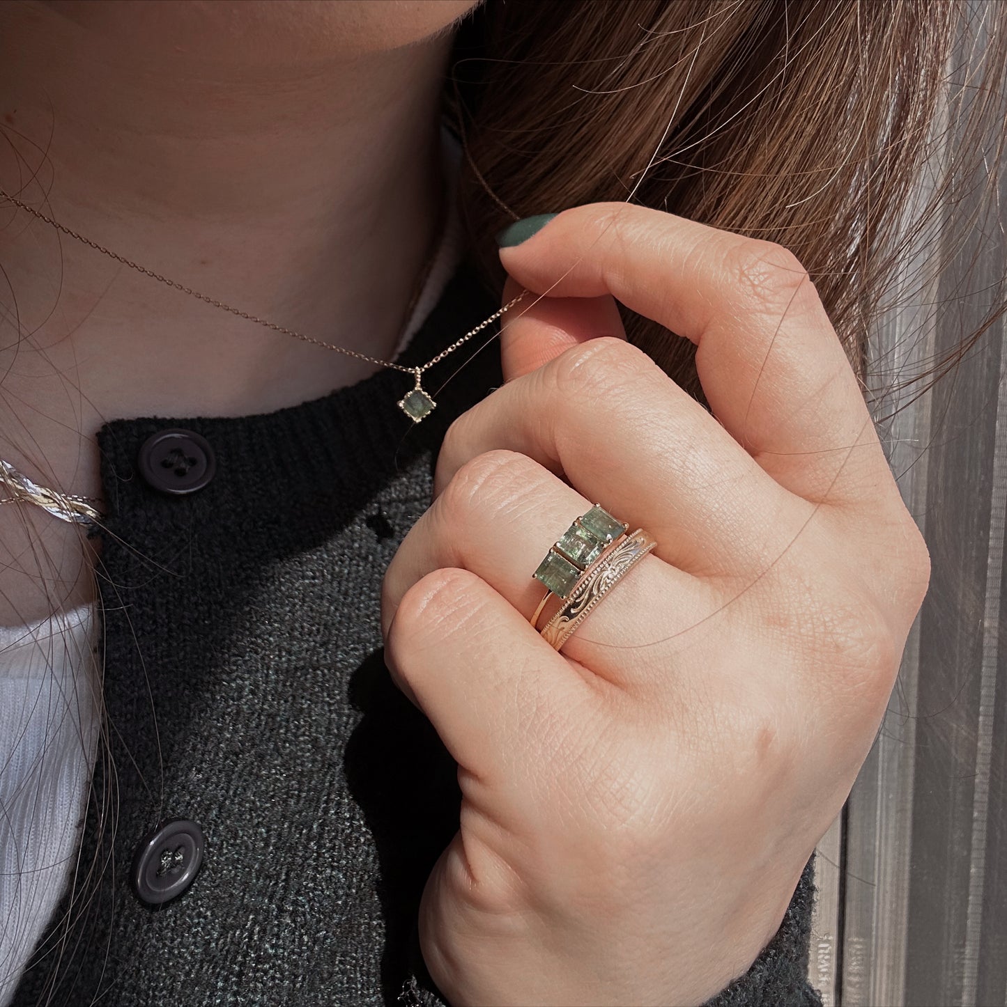A close-up of a hand wearing the Solid Gold Filigree Ring and holding a necklace with a green gemstone pendant. The person wears a dark cardigan with black buttons, and sunlight illuminates their hair and jewelry.