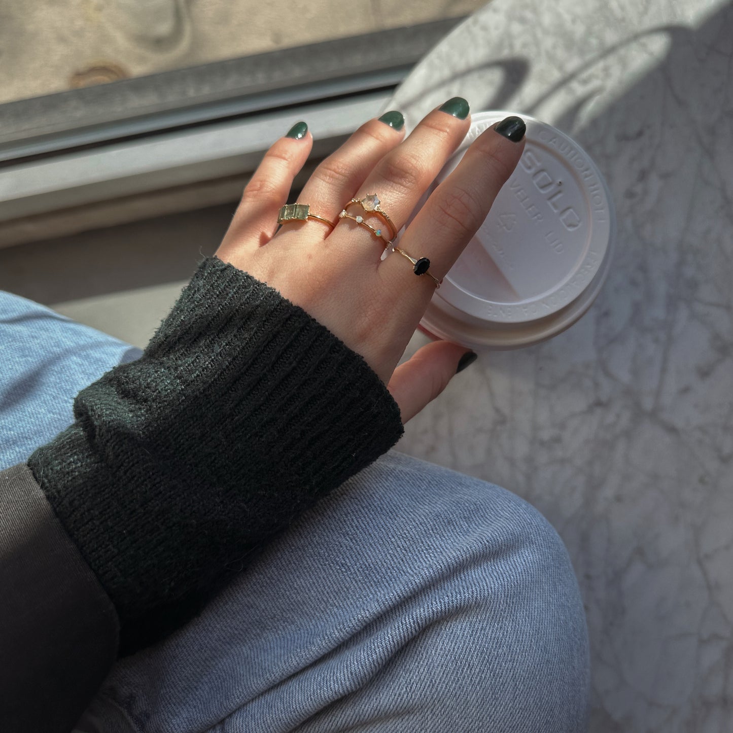 A hand with dark green nail polish and several rings, including the Solid Gold Mystic Labradorite Hexagon Ring for Protection, holds a white coffee cup. The person wears a dark sweater and light blue jeans by a sunlit marble windowsill.