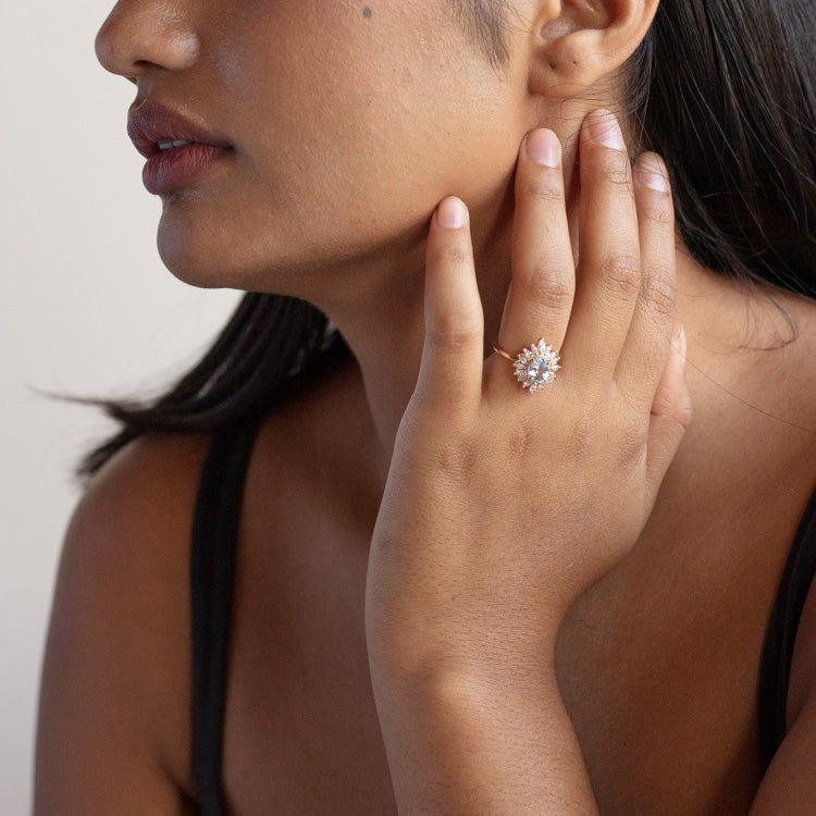 A woman with medium brown skin and dark hair touches her face, showcasing the Lana Ring with Aquamarine & CZ—a 14k solid gold piece featuring a round, sunburst-style cluster of sparkling stones. She wears a black sleeveless top.
