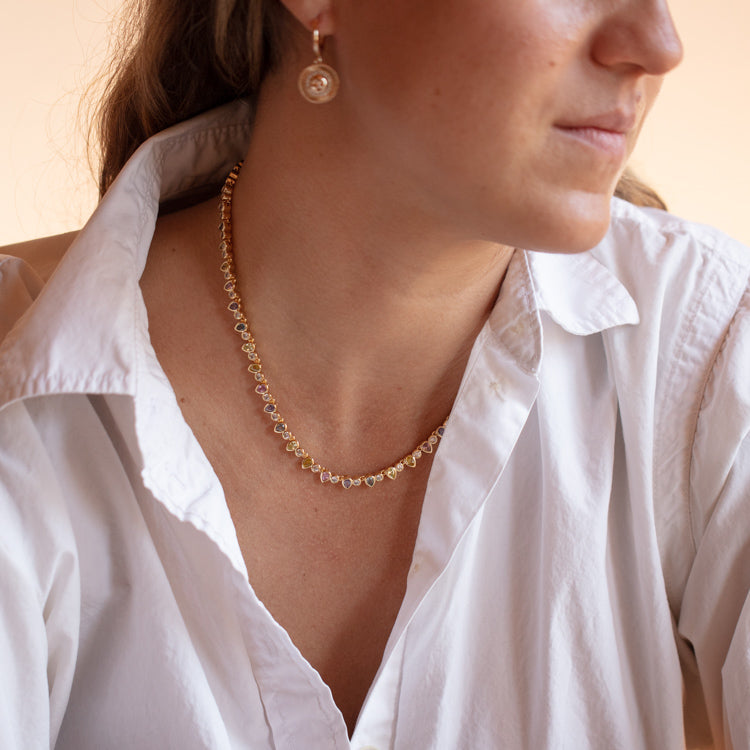 A woman in a white collared shirt wears the Teardrop Tennis Necklace, a delicate 14k gold piece with small charms, along with a matching earring. She is shown from the shoulders up in soft lighting, her face partly turned to the side.