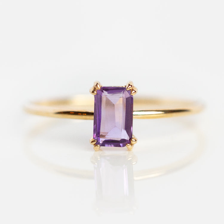The Solid Gold Essential Baguette Birthstone Ring features a rectangular faceted purple gemstone in a four-prong setting, showcased on a white background.