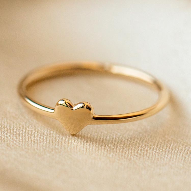 The Solid Gold Engravable Heart Ring Sample Size 9 is a delicate 9k yellow gold ring featuring a small heart-shaped design atop a 1.2 mm band, shown resting on soft beige fabric.