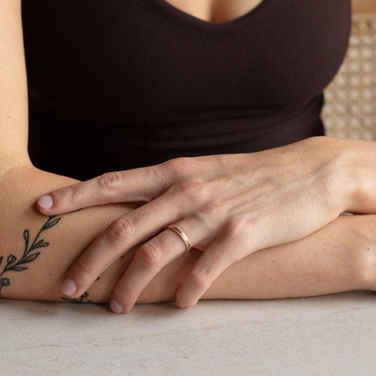A person with a leafy branch tattoo on their forearm rests their hands on a table, wearing the Solid Gold Filigree Ring on their finger and dressed in a sleeveless, dark top.