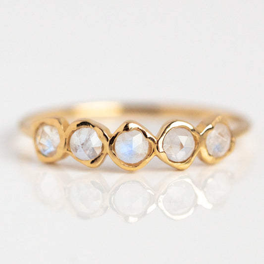 The Solid Gold Organic Moonstone Ring features five round faceted moonstones in scalloped bezels, arranged in a row and displayed on a white background with a subtle reflection.