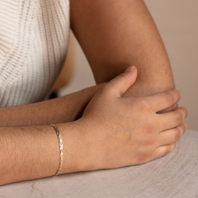 A close-up of a person resting their arms on a light surface, wearing a Solid Gold Mixed Metal Braided Bracelet and a sleeveless, ribbed white top.