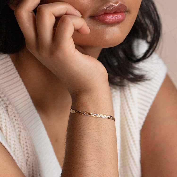 A woman in a sleeveless white knit top rests her chin on her hand, highlighting the Solid Gold Mixed Metal Braided Bracelet on her wrist. Only the lower part of her face and upper body are visible.