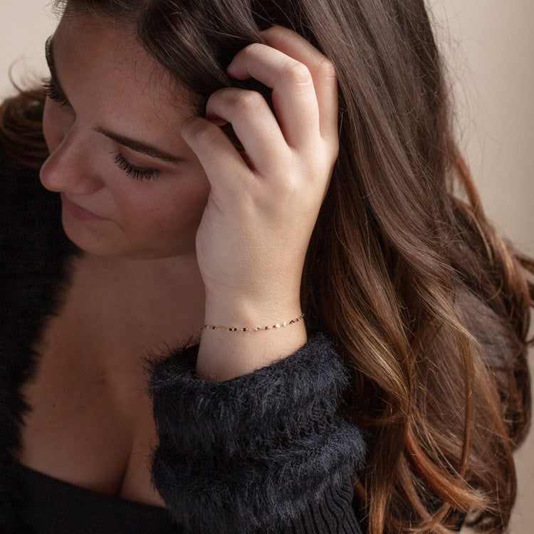 A woman with long brown hair in a fuzzy black sweater looks down, gently touching her hair. On her wrist is the Solid Gold Bold Sparkle Link Chain Bracelet, crafted from 14k yellow gold for an elegant touch.