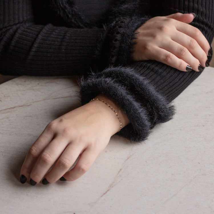 A person with black nail polish rests their hands on a light marble surface, wearing a black ribbed sweater with fuzzy cuffs and the Solid Gold Bold Sparkle Link Chain Bracelet.
