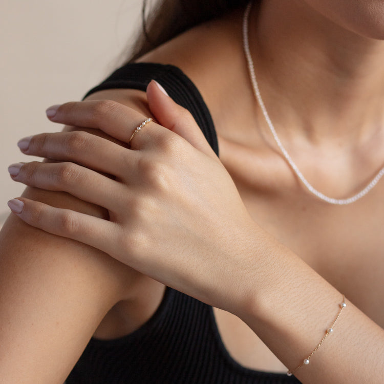 A woman in a black sleeveless top showcases the Solid Gold Simple Pearl Chain Bracelet, featuring delicate freshwater pearls. She pairs it with a thin gold chain, a simple ring, and pale pink manicured nails.