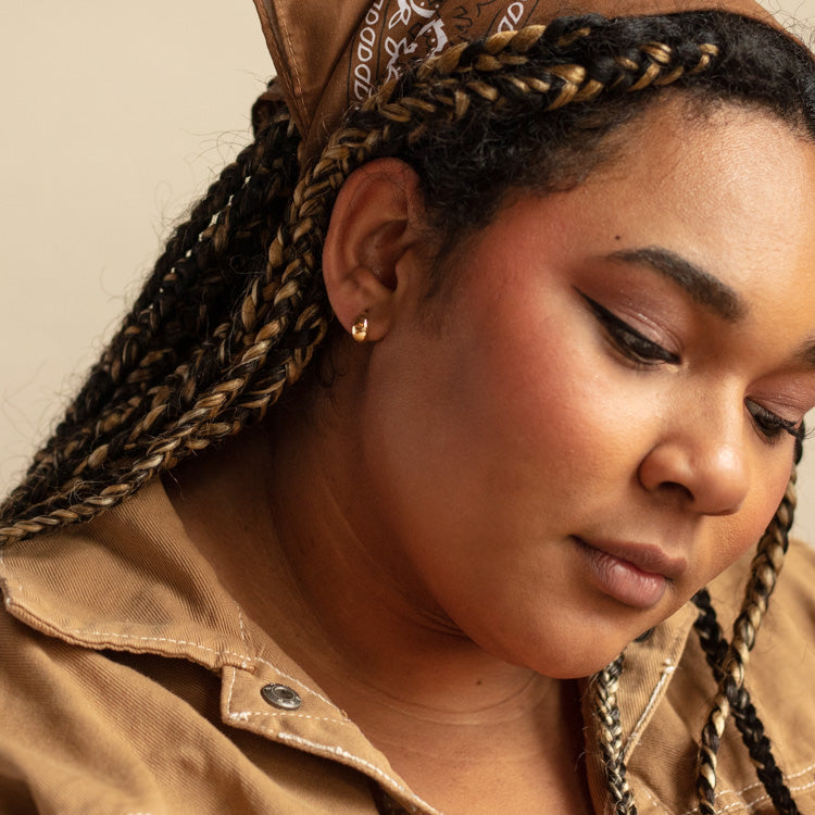 A woman with braided hair and a brown patterned headscarf looks down thoughtfully. She wears a tan jacket and Solid Gold Chunky Huggie Hoops, with natural makeup and soft lighting highlighting her face.