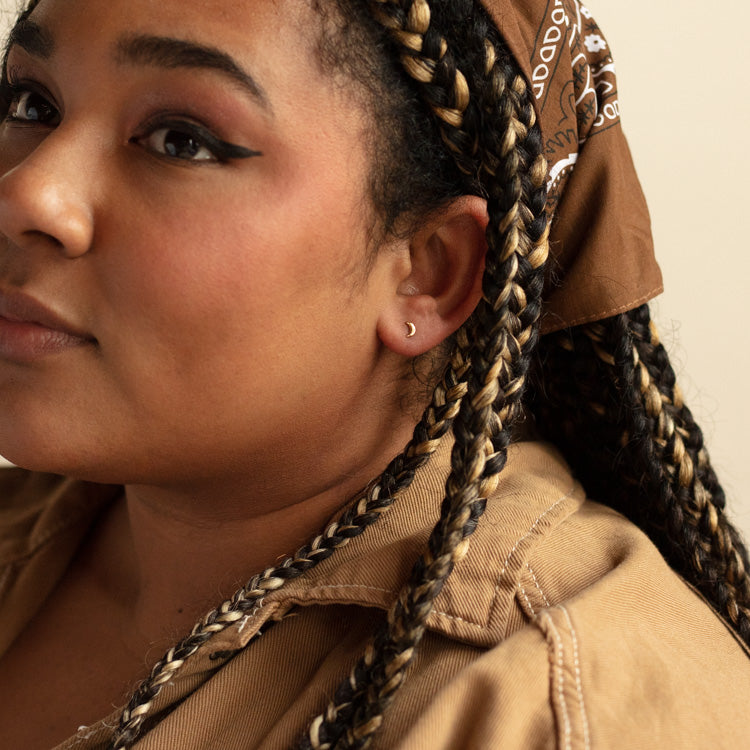 A woman with medium skin tone, long braided hair, and subtle makeup wears a brown bandana, tan top, and a dainty Solid Gold Moon Earring. Only part of her face is shown.