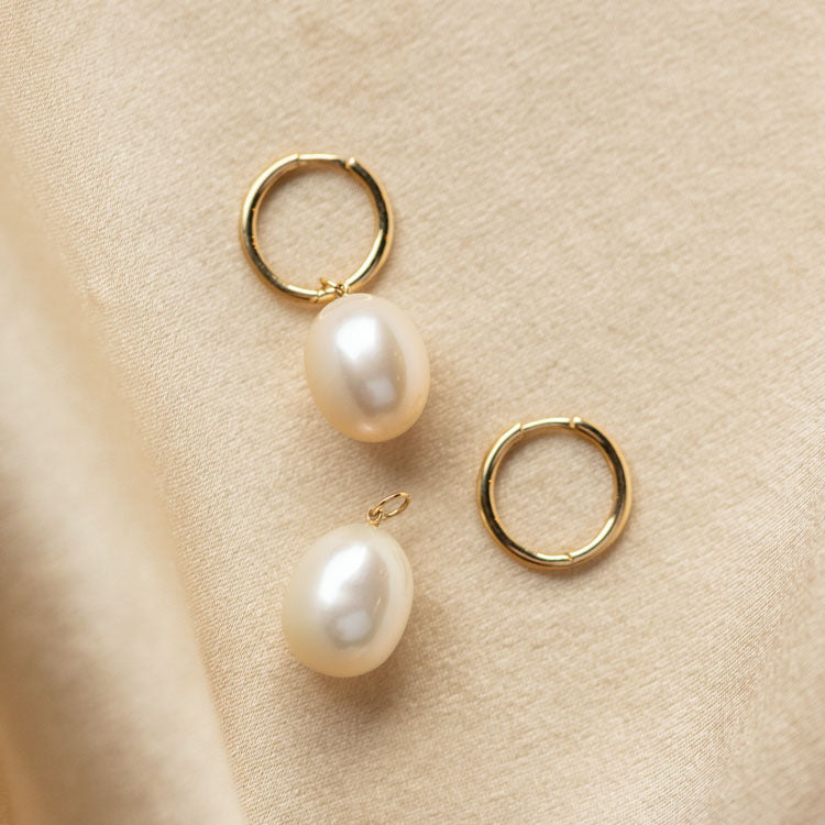 The Solid Gold Elegant Pearl Charm Huggies, featuring a removable pearl charm, rest on a soft beige fabric background.