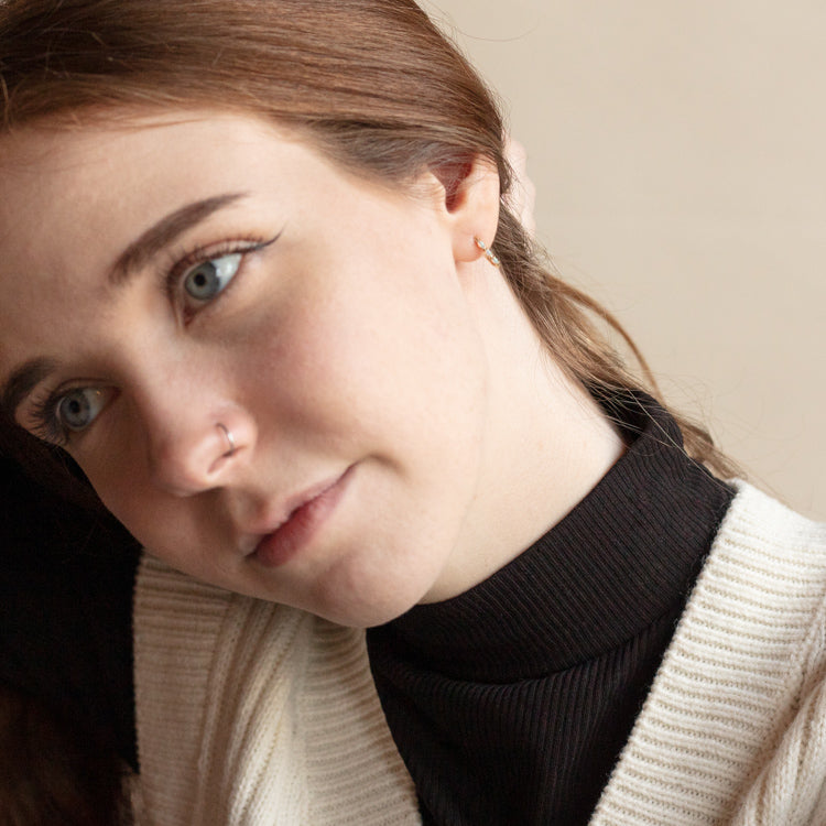 A young woman with fair skin and blue eyes wears a black turtleneck and cream sweater. She gazes thoughtfully to the side, her head slightly tilted, wearing the Solid Gold Opal Oblong Huggie Hoops Sample.